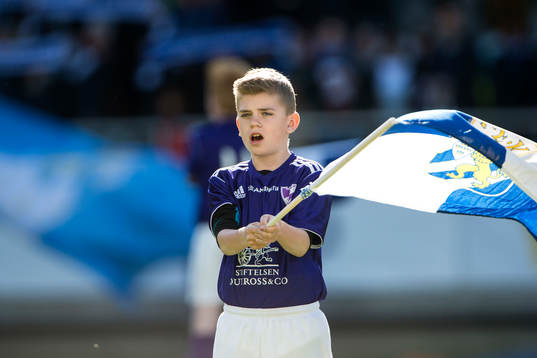En maskot med en blåvit flagga  innan fotbollsmatchen i