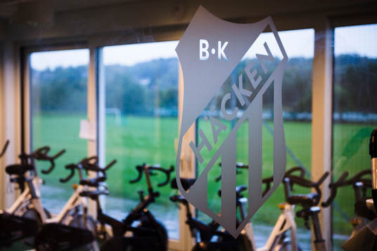 Häckens klubbmärke framför ett rum med motionscyklar på