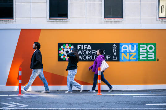 Three persons walk past a board with the logo for FIFA